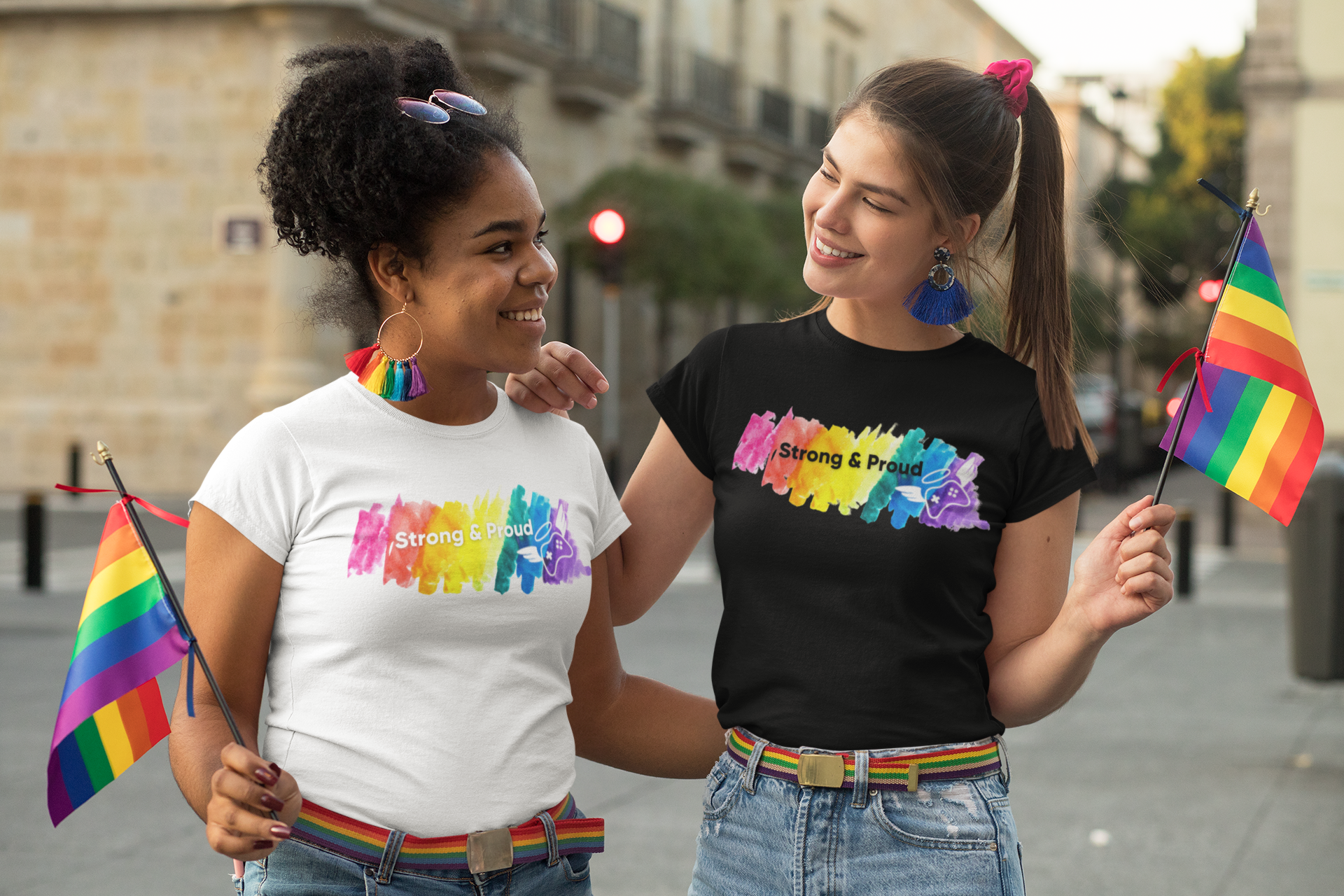 two queer femme-presenting people holding rainbow flags and smiling at each other during Pride month wearing Extra Life Pride shirts