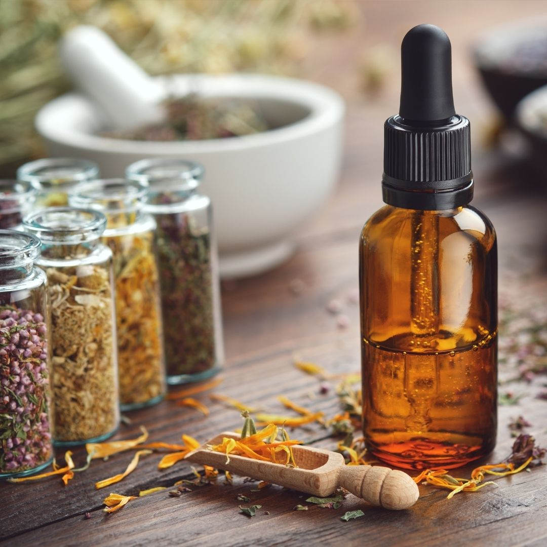 amber dropper vial half filled with liquid next to small glass vials filled with various dried herbs and a mortar and pestle in the background