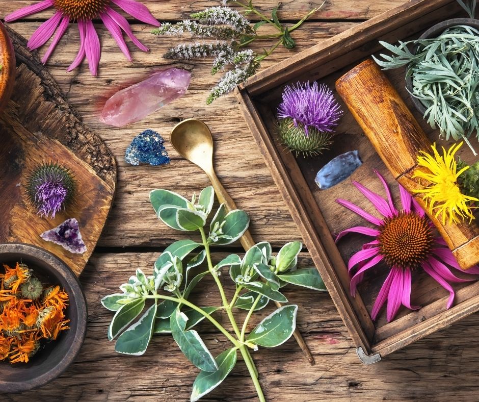 table shot of various herbs, flowers, and crystals scattered around as if in the middle of crafting
